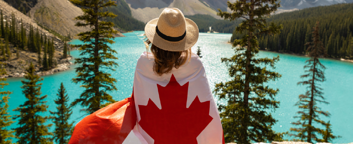 Banner Canadá: Mulher de costas com chapéu na cabeça, vestindo a bandeira do Canadá contemplando um lago de águas cristalinas.