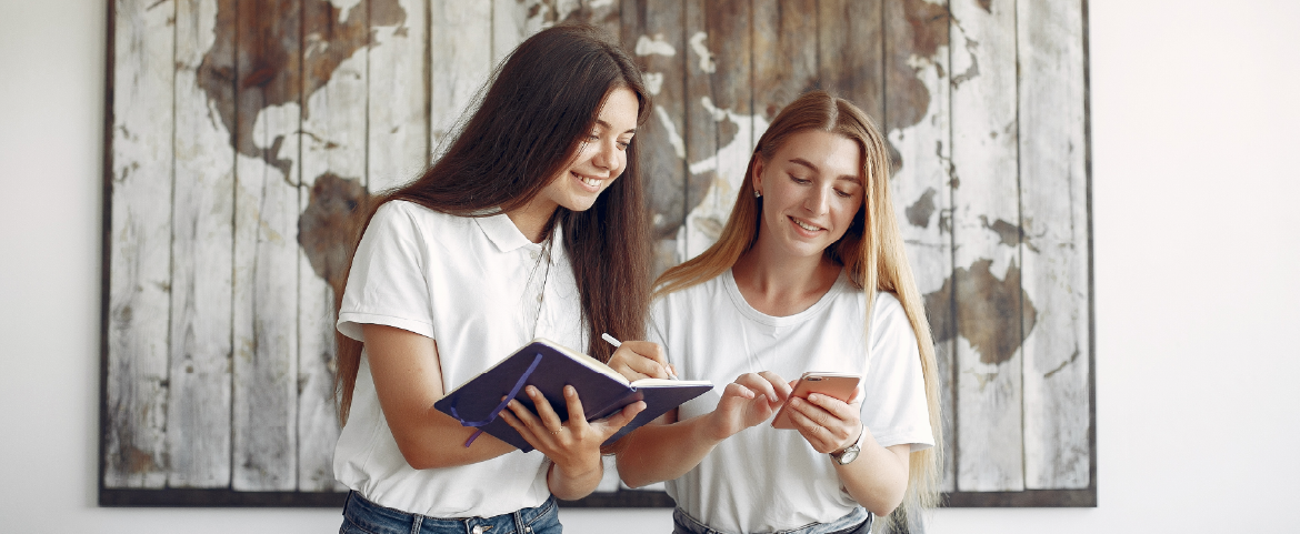 Duas mulheres em pé segurando livros e um mapa mundi ao fundo Duas mulheres em pé segurando livro e celular e um mapa mundi ao fundo