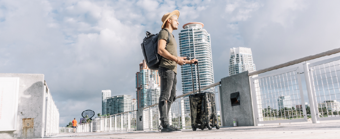 Homem de chapéu,, mochila nas costas e bagagem de bordo fazendo turismo em uma metrópole.