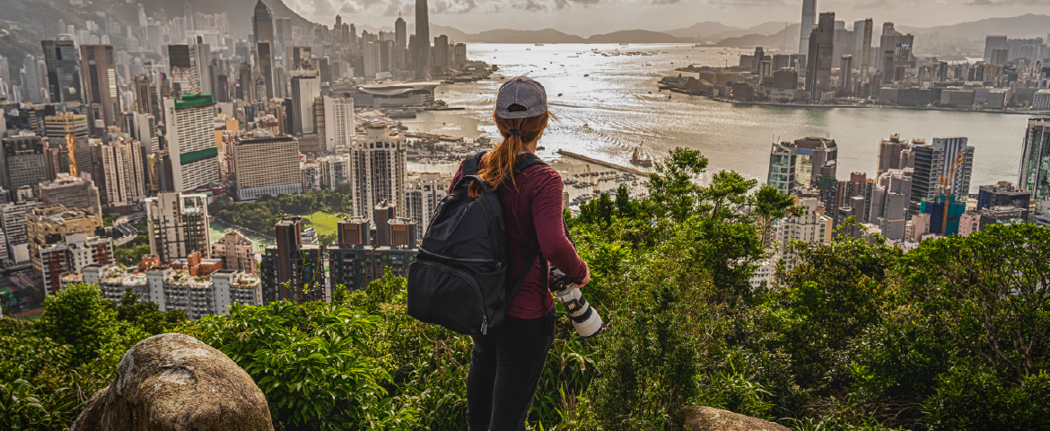 Garota ruiva no alto de uma montanha com mochila nas costas e câmera fotográfica na mão.