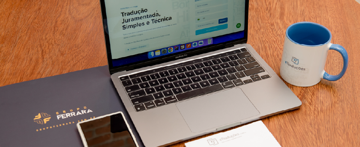 Laptop com site da eTraduções aberto em cima de mesa de madeira com pasta do Grupo Ferrara e caneca da eTraduções.
